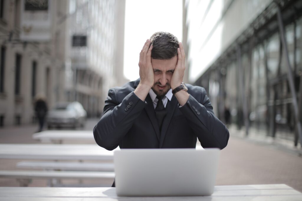 A businessman in a suit looks stressed, sitting outdoors with a laptop in a city environment.