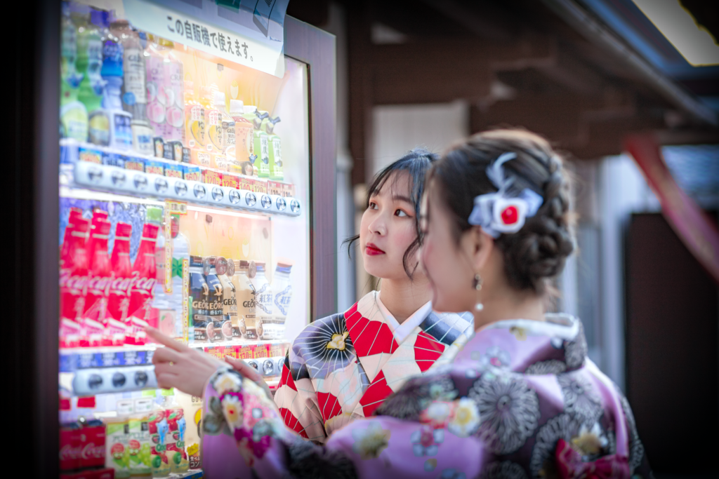 無人ビジネスとは? Two women wearing kimonos choose drinks from an outdoor vending machine during the day.