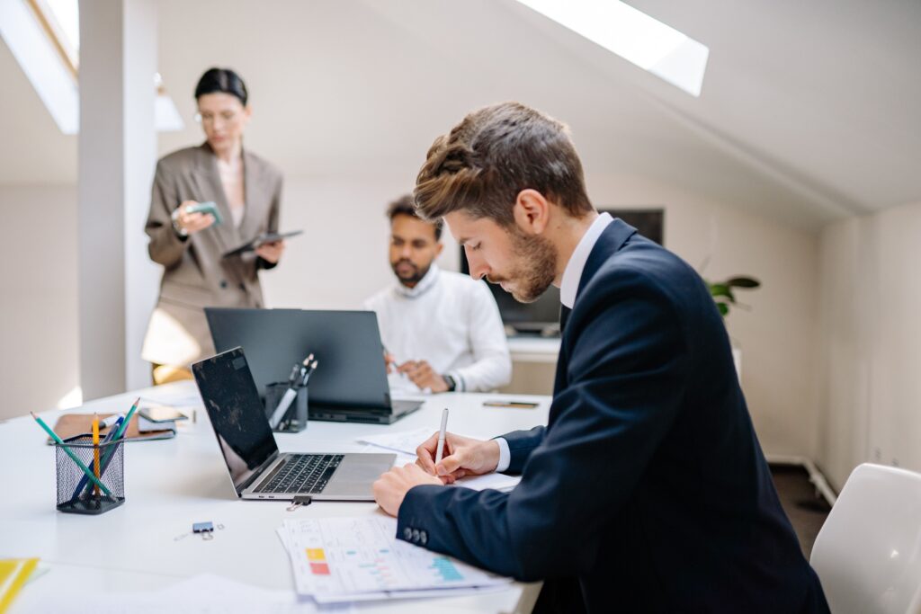 100万円以下でできる無人ビジネス一覧 Three colleagues working on laptops and documents in a bright office setting.