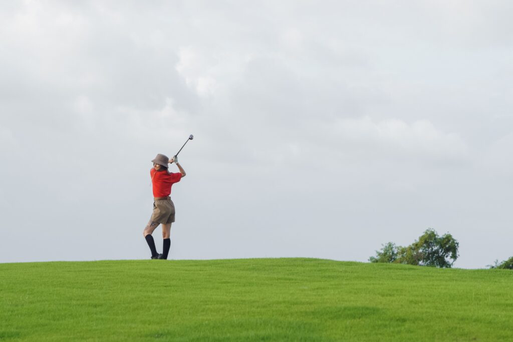 【保存版】無人インドアゴルフ開業ロードマップ!準備からオープンまでの全5ステップ A woman in red shirt swinging a golf club on a lush green course under a cloudy sky.